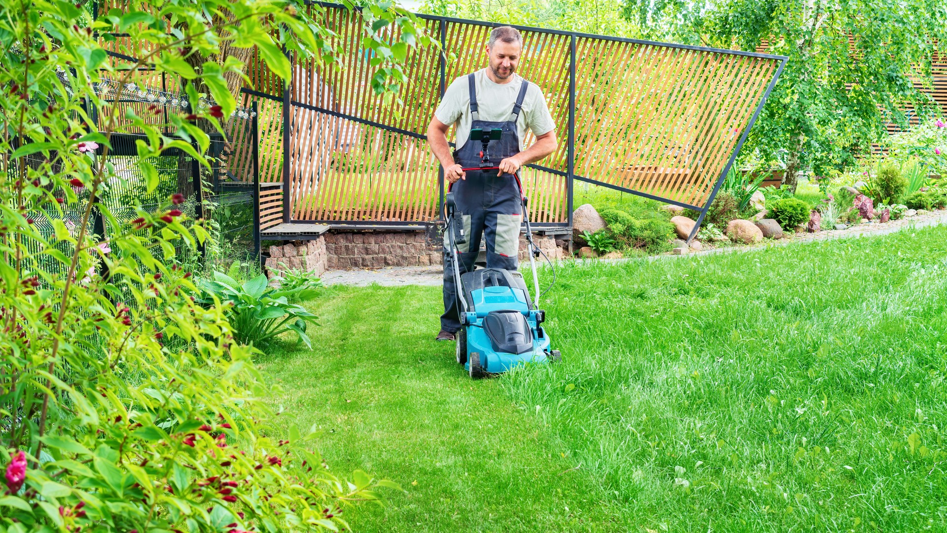 A man mows the grass with an electric lawn mower on a hot summer day. Gardener with a lawn mower in a beautiful landscape design. A hired worker tends the garden and does housework. Gardener services.